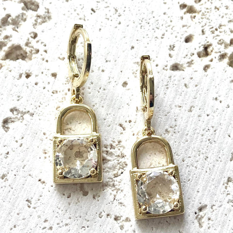 A pair of gold lock earrings with clear crystal accents on a white textured surface.