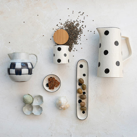 Left to right: a small jar with checkboards, an egg holder with one egg, a garlic, a small dish with food, a coffee container with coffee around it, a long plate with olives, and a large jar with dots.