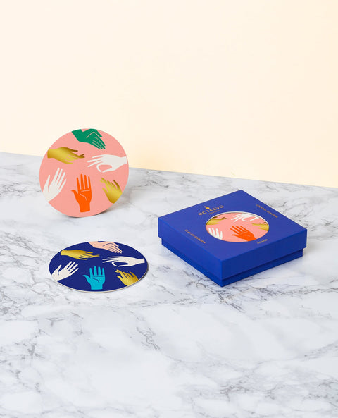 Two coasters, one pink, one blue, with printed hands doing Hamsa signs and the blue box.