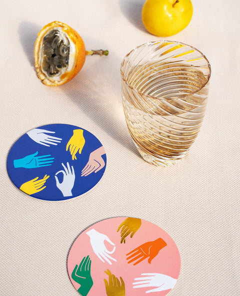 Two coasters, one pink, one blue, with printed hands doing Hamsa signs and a glass of water with some fruits around.