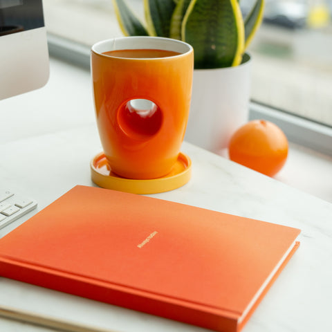 Orange mug with a unique design on a white surface next to an orange notebook and a potted plant.