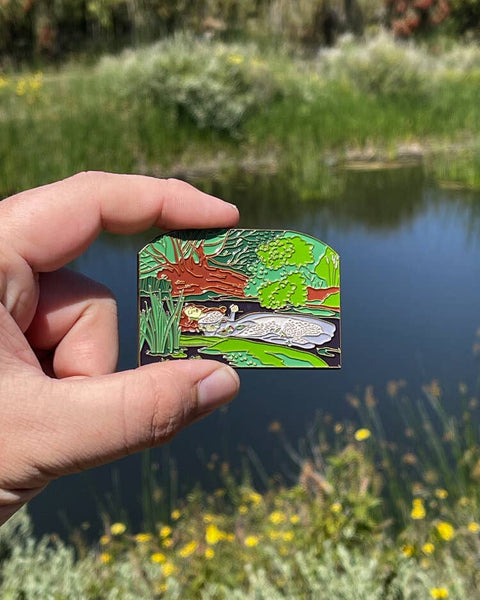 Hand holding a nature-themed pin with a pond and fish in the foreground, blurred natural background