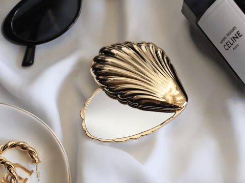 Gold shell-shaped mirror on a white fabric background with Celine handbag partially visible.