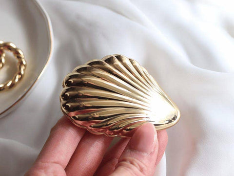 Gold shell-shaped mirror held by a hand on a white fabric background