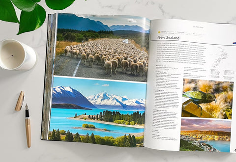 Open book showing scenic images of New Zealand with a cup and pen on a table.