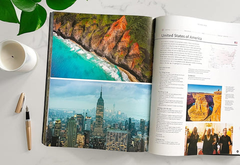 Open book showing images of a beach, cityscape, and people on a white surface with a cup and pen.