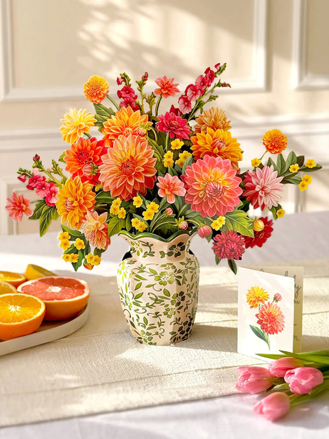 Paper bouquet of colorful flowers in a decorative vase on a table with fruits and a card.