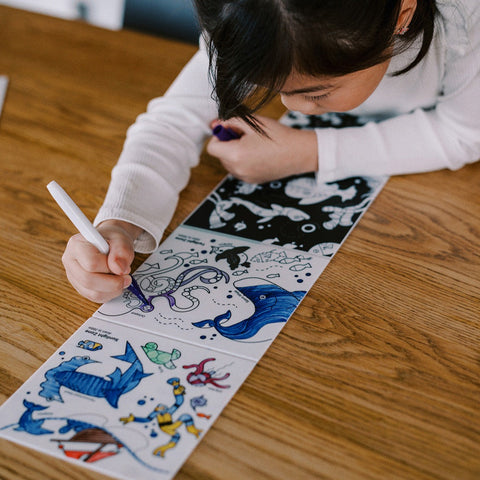 Child coloring a book of marine life with markers on a wooden table.
