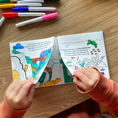 Coloring book with open pages on a wooden table, child's hands holding markers.