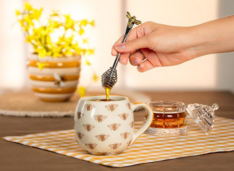 Hand pouring honey from a spoon into a mug with bee patterns on a wooden table.