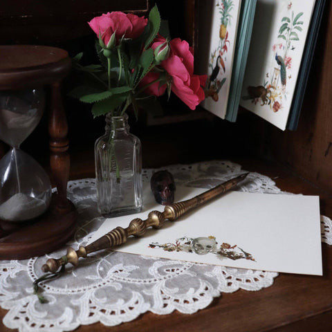 Vintage-style setup with notecards, a bottle of pink roses, a quill pen, and an hourglass on a lace tablecloth.