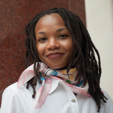 Woman with dreadlocks wearing a colorful scarf and white jacket against a red and beige wall.
