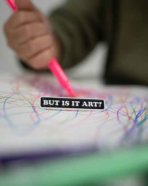Child's hand with a pink crayon on a piece of paper with colorful lines, overlaid with a black pin reading 'BUT IS IT ART?'.