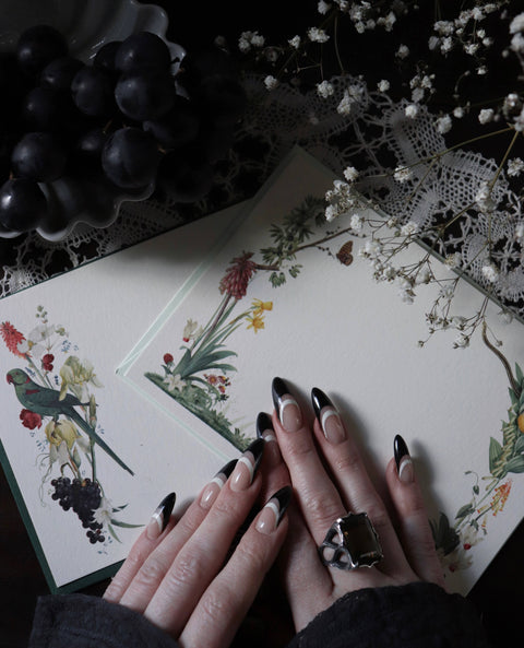 Hands with black nail polish on a floral card with grapes and dried flowers in the background
