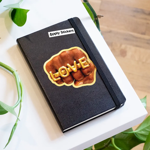 Black notebook with a 'LOVE' sticker on a white surface with green leaves in the background