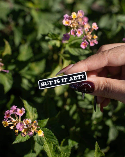 Hand holding a pin with 'BUT IS IT ART?' text against a floral background