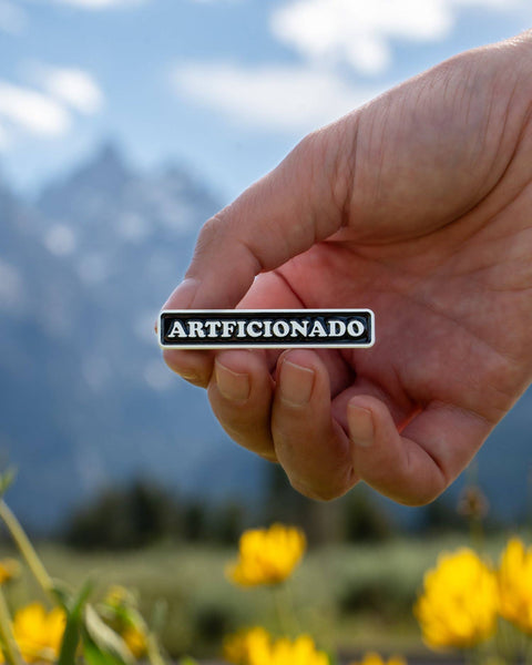 Hand holding a 'Artficionado' pin with a mountain and yellow flowers in the background