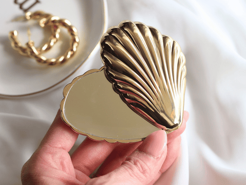 Gold shell-shaped compact mirror held by a hand with a blurred background