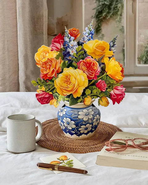 Colorful paper flower arrangement in a decorative vase on a table with a mug, pen, and book.